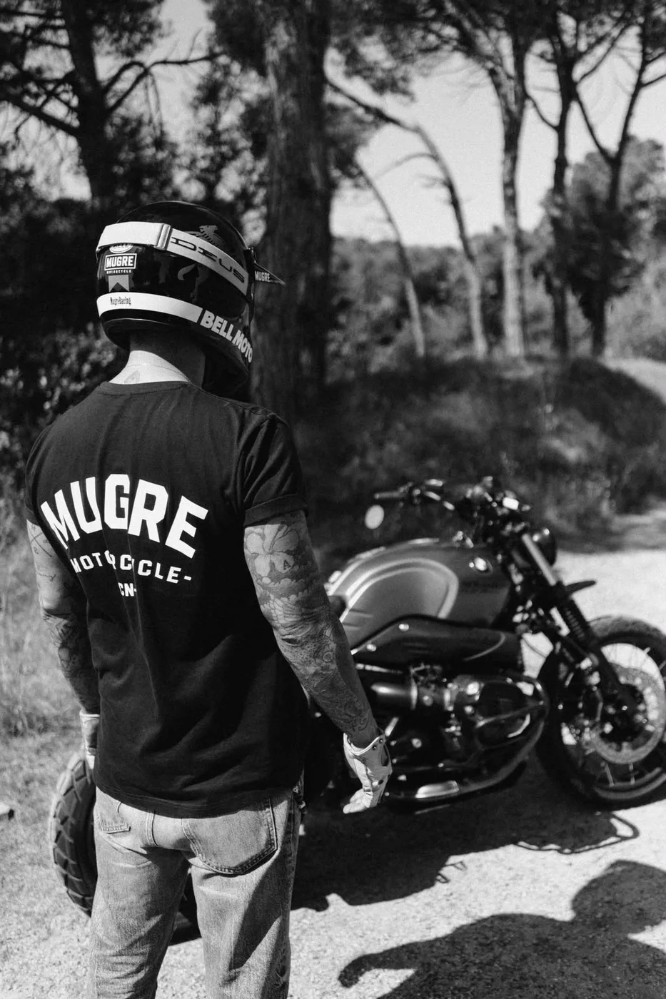 Rider con camiseta Mugre Motorcycle junto a una moto en carretera forestal