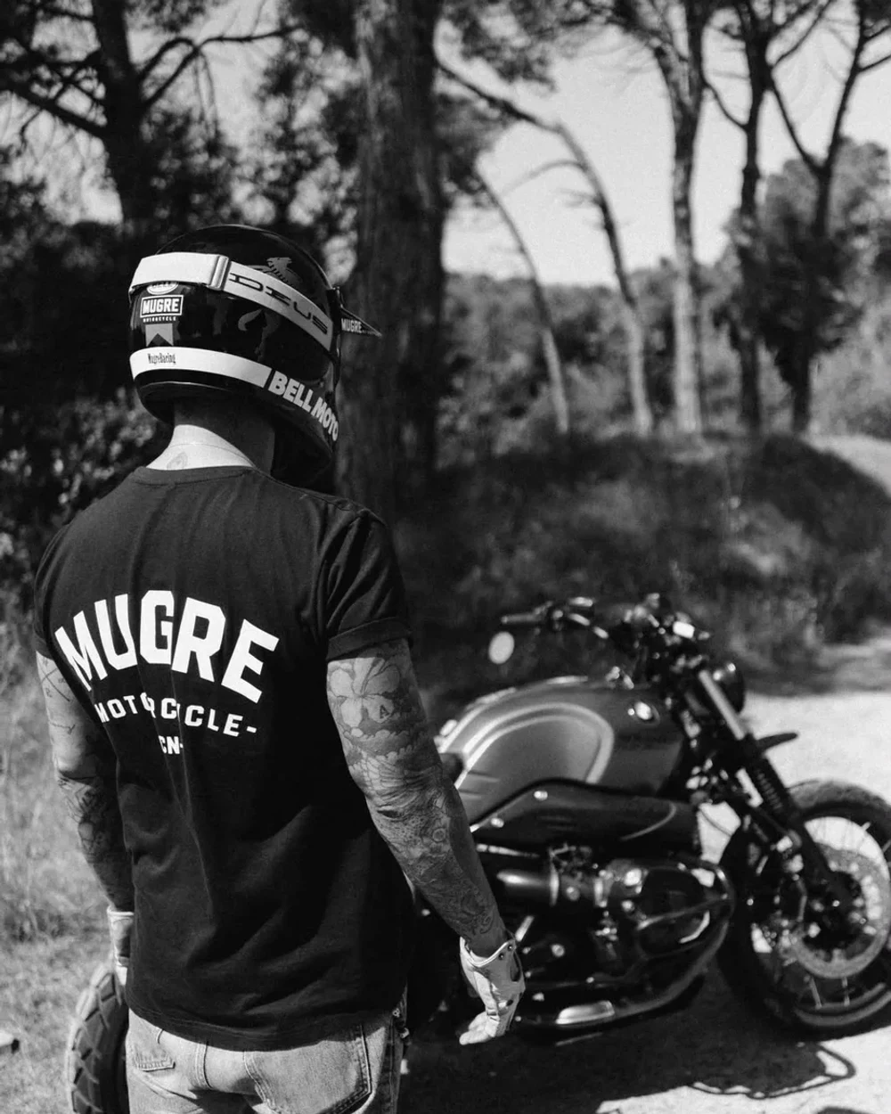 Rider con la camiseta BARCELONA de espaldas, junto a su moto en una carretera forestal